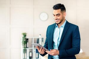 A young Middle Eastern man in a suit is engaged with a tablet in a contemporary office. He appears focused and cheerful while working, reflecting a dynamic work environment. photo