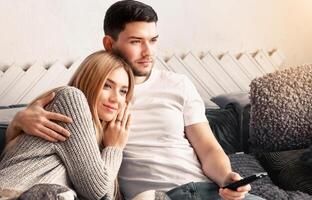 A young couple nestles on a cozy couch, with the woman resting her head on the man's shoulder as they enjoy watching a show together. Soft light filters through the space, creating a warm atmosphere. photo