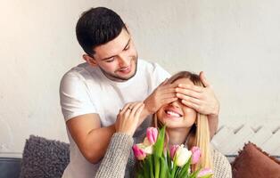 A man joyfully surprises a smiling woman with a bouquet of flowers while playfully covering her eyes. They share a warm and intimate moment in a cozy indoor setting. photo