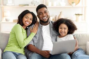 A smiling black family of three gathers on a couch in their cozy living room, engaging with a laptop. They appear joyful, reflecting a warm and loving atmosphere during family time. photo
