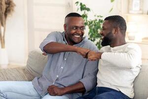Two men are sitting on a couch in a bright room, laughing and sharing a fist bump. They express joy and camaraderie, showcasing a strong bond during a relaxed moment together. photo