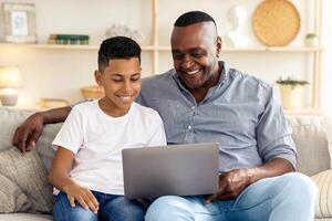 A father and son engage in a delightful moment, sharing smiles and laughter while exploring content on a laptop in a cozy living room on a weekend afternoon. photo
