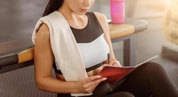 A gym-goer relaxes after a workout, taking notes in a notebook while sipping on a colorful beverage. She reflects on her fitness routine and goals in a bright and motivating environment. photo
