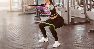 A woman is engaged in a squat exercise using a resistance band in a well-lit gym. She focuses on maintaining proper form while working out during a morning fitness session. photo