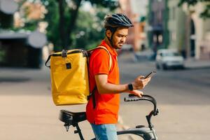 Modern mobile applications. Young man in safety helmet, with big yellow bag, on bicycle looks at smartphone, outdoors, profile, copy space photo
