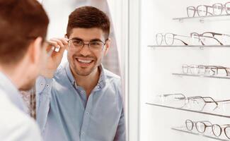 A young man examines himself in the mirror while trying on a stylish pair of glasses in an eyewear store, displaying a joyful expression as he checks the fit and style. photo