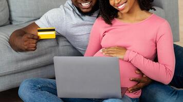 Positive black pregnant couple shopping online at home, getting ready for parenthood, using laptop and credit card, empty space photo