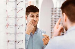 A young man stands in front of a mirror, applying contact lens solution to his eye while surrounded by various eyeglasses in a retail store dedicated to eyewear. photo