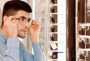 A young man inspects his appearance while trying on various glasses in an eyewear shop, focusing intently on finding the perfect pair. The setting is bright and inviting, showcasing several options. photo