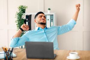 A young Arab businessman stretches at his office desk, showing a moment of relaxation during a busy workday. His laptop sits open while he enjoys a brief break from tasks. photo