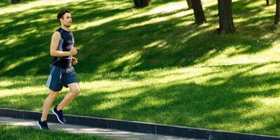 Physical activity outside. Guy in sportswear with sneakers and fitness tracker runs at path in park, free space, sun flare photo