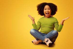 Joyful little girl sitting on floor shouting and raising hands with excitement, posing over yellow studio background, empty space photo