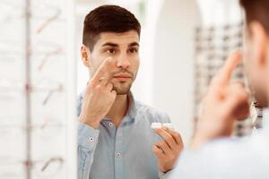 A man stands in a store, carefully applying contact lenses as he looks into a mirror. He holds the lens case in one hand, focused on his reflection. photo