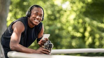 Smiling african sportsman drinking water and listening to music with smartphone and wireless headset while resting, exercising at park, free space photo