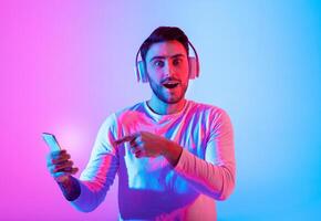 A man with headphones smiles while pointing at his smartphone illuminated by colorful lights. His expression reflects excitement and engagement with the music he is enjoying. photo