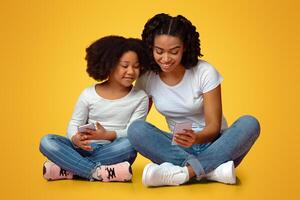 Smiling Black Mother Teaching Her Little Daughter How To Use Smartphone While Sitting Together On Floor Over Pink Studio Background, Copy Space photo