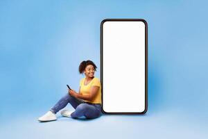 A cheerful African American woman sits on the floor with her smartphone, smiling at the camera. She is casually dressed in a yellow top and jeans, with a large blank smartphone screen beside her. photo