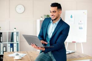 A young Middle Eastern entrepreneur sits at a desk in a contemporary office, smiling as he works on his laptop. The workspace features organized files, a clock, and a whiteboard. photo