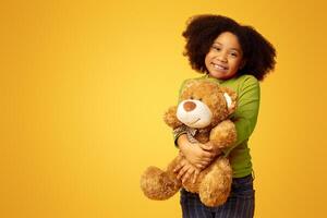 Portrait of adorable little black girl with teddy bear in hands standing over yellow background, posing and smiling to camera, free space photo