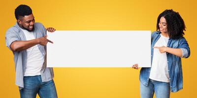 A happy couple stands side by side, pointing at a large blank sign. They are smiling and wearing casual clothing, showcasing their enthusiasm against a vibrant yellow backdrop. photo