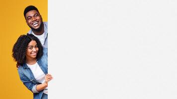 Two joyful young adults peek from behind a blank space, sharing a light-hearted moment. The vibrant yellow background adds to the fun atmosphere of their interaction. photo