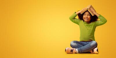 Education Concept. Lovely little black child sitting on floor and wearing book as a hat, having fun over yellow background photo