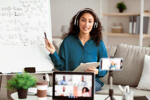 A young Middle Eastern woman with brunette hair is teaching an online math class. She is using a whiteboard to explain concepts, while students engage through calls. photo