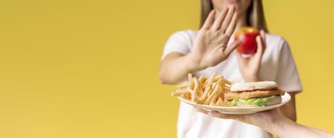 A person stands against a vibrant yellow background, refusing a plate of fries and a sandwich, while confidently holding an apple. This represents a commitment to healthier eating choices, copy space photo