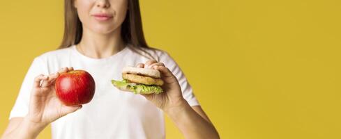 A woman stands confidently, holding an apple in one hand and a sandwich in the other, symbolizing the decision between healthy and indulgent food choices, copy space photo