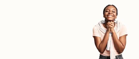 A cheerful young woman stands with her hands clasped together, smiling broadly in a bright, minimalistic environment. Her joyful expression conveys feelings of anticipation and gratitude. photo