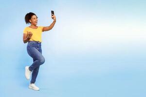 A cheerful African American woman stands against a light blue background, smiling as she captures a selfie with her smartphone while wearing casual attire and stylish sneakers, copy space photo