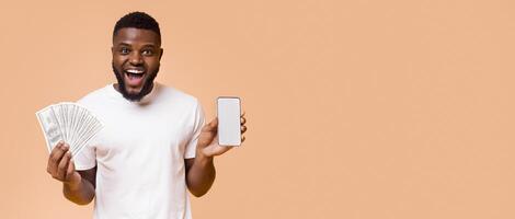 Making money on internet. Happy african american guy showing showing cellphone with blank screen and bunch of money, orange background, free space photo