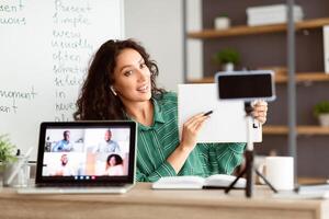 A brunette woman leads an online class from a cozy room, showcasing teaching materials on her tablet while explaining concepts to attentive students. The scene reflects effective remote learning. photo