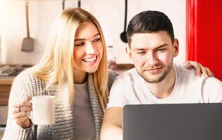 A couple shares a joyful moment in a bright kitchen, sipping coffee as they look at a laptop screen together, smiling and engaging in conversation. photo