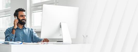 Handsome indian man office manager talking on cellphone at his workplace in office, sitting in front of computer, enjoying pleasant phone conversation, full length shot photo