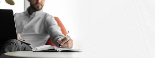Close up of focused business man sitting at desk working using pc taking notes in notebook at office, selective focus on hand holding pen in foreground, blurred background. Studying online photo