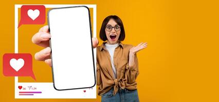 A young influencer with glasses excitedly shows a smartphone displaying a blank screen while interacting with social media. The backdrop is bright yellow, creating a cheerful atmosphere. photo