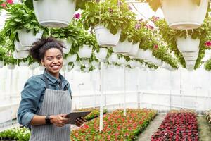 Gardener using digital tablet in greenhouse. Smiling african american girl with smart watch and tablet, in orangery with flowers hanging from ceiling, free space photo