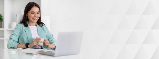 Glad millennial caucasian lady in suit resting at workplace with cup of tea and computer in office interior. Relax, coffee break, business with device, manager work, call and meeting photo