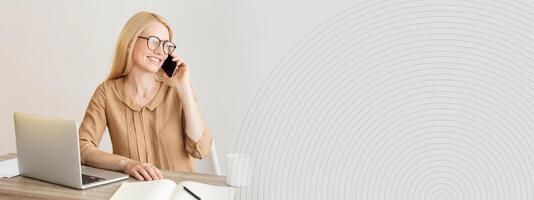 A woman in a beige blouse smiles while conversing on her phone. She is seated at a wooden desk with a laptop, an open notebook, and a plant in a bright, minimalistic office environment, copy space photo