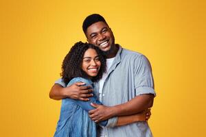 A happy couple stands closely together, embracing with smiles on their faces. The warm yellow backdrop highlights their joyful expressions and connection, capturing a moment of love. photo