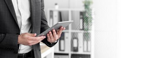 A business professional in a suit is engaged with a tablet, standing in an office space featuring modern decor and organized shelves, focusing on technology in daily work. photo
