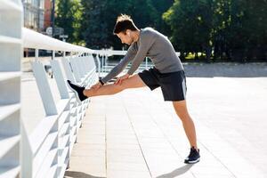 Stretching and end of workout. Muscular guy in sportswear puts his foot on railing and does exercises in sunlight on street, side view, free space photo