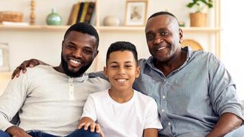 A grandfather, father, and son sit closely together on a couch, smiling warmly at the camera. The cozy living room features light decor and a sense of togetherness. photo