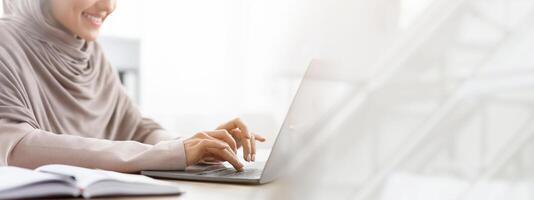 Unrecognizable muslim woman in hijab working on laptop computer in office, typing on keyboard and smiling, cropped image with empty space photo