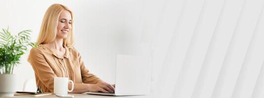 A woman is focused on her laptop in a bright, modern workspace adorned with greenery. She enjoys her task with a warm smile, accompanied by a steaming mug beside her, copy space photo