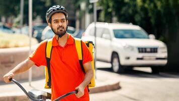Mobile work in city, fast delivery and sporty lifestyle. Attractive millennial arabian guy courier in orange t-shirt and protective helmet, with big backpack and bicycle looks for address of client photo