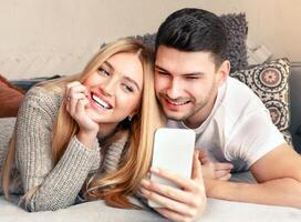 A young couple relaxes on a soft couch, smiling as they share a light-hearted moment while looking at their smartphone. They are dressed casually, enjoying each other's company. photo