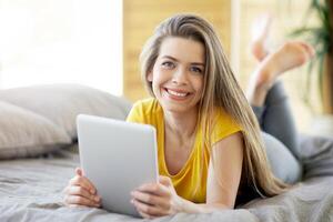 Smiling millennial woman using tablet computer for online work or education on bed at home, copy space photo