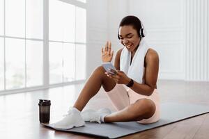 A young African American woman engages in fitness at home, listening to music while smiling and checking her smartphone. She is in a bright room, showcasing a healthy lifestyle. photo